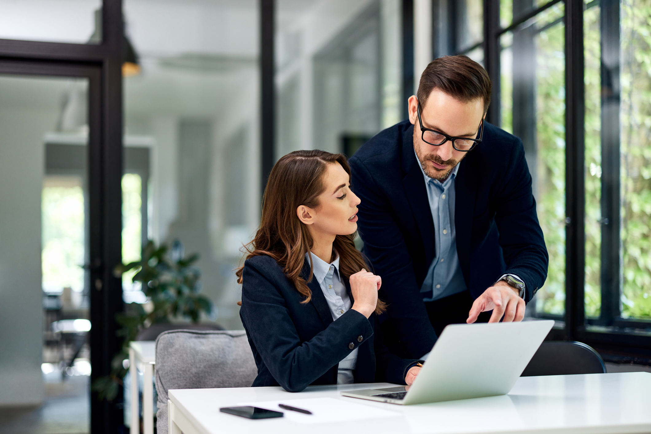 A man and woman in professional attire discuss work over a laptop in an office setting