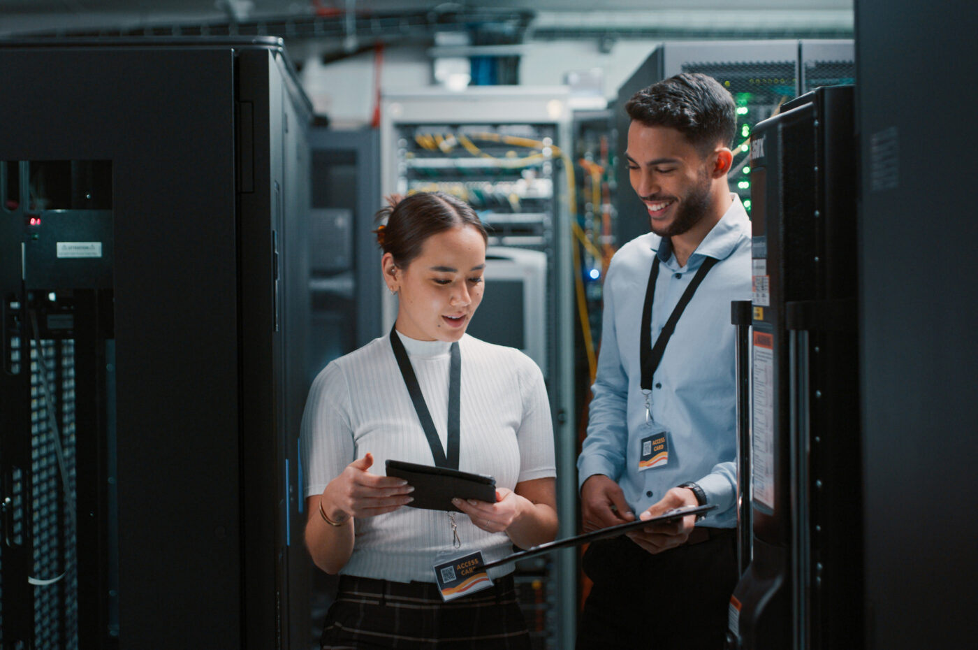 Two individuals discussing in a server room, holding tablets and smiling