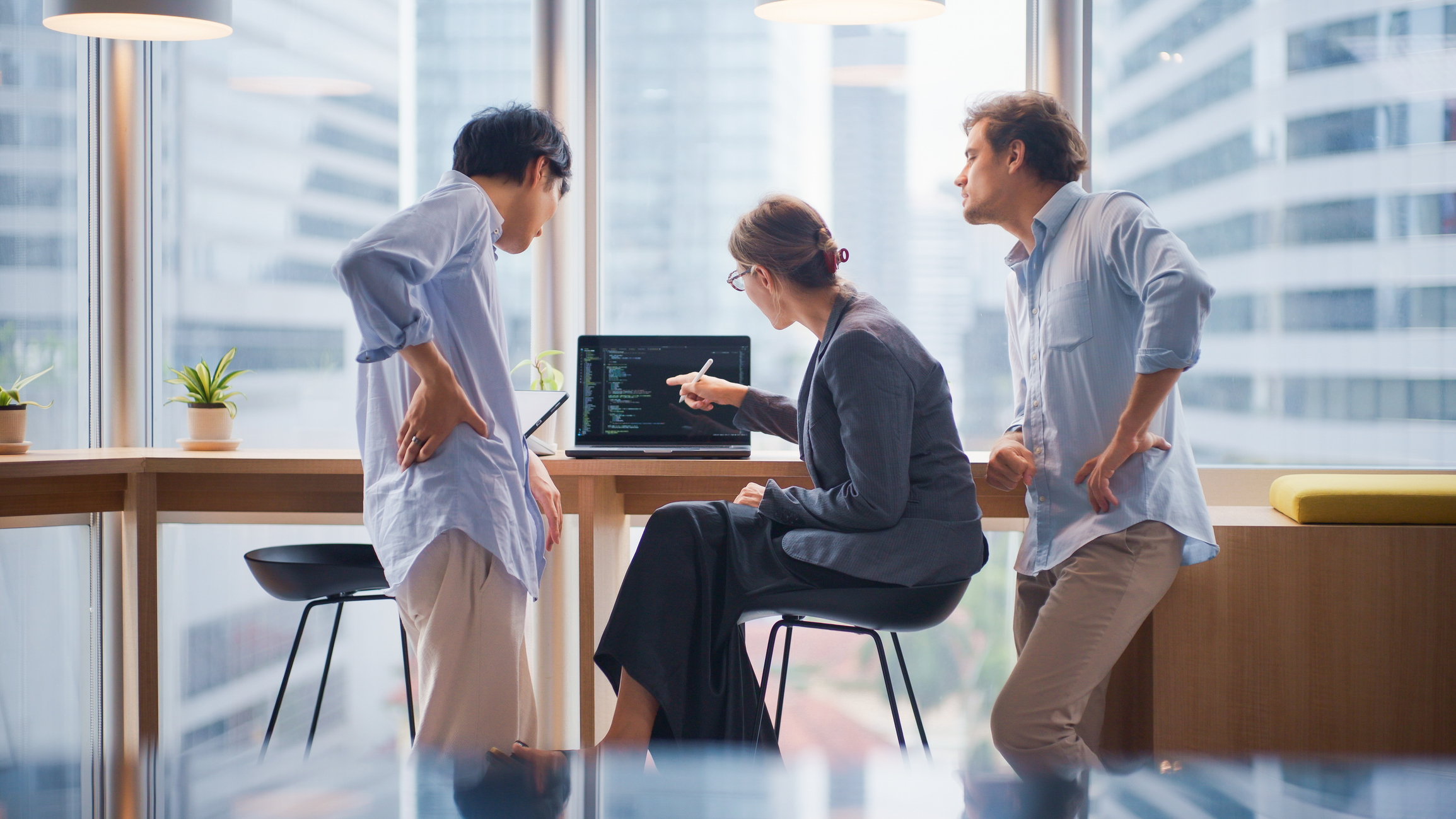 IStock-2231487646.jpg, three colleagues discussing code on laptop in office with large windows