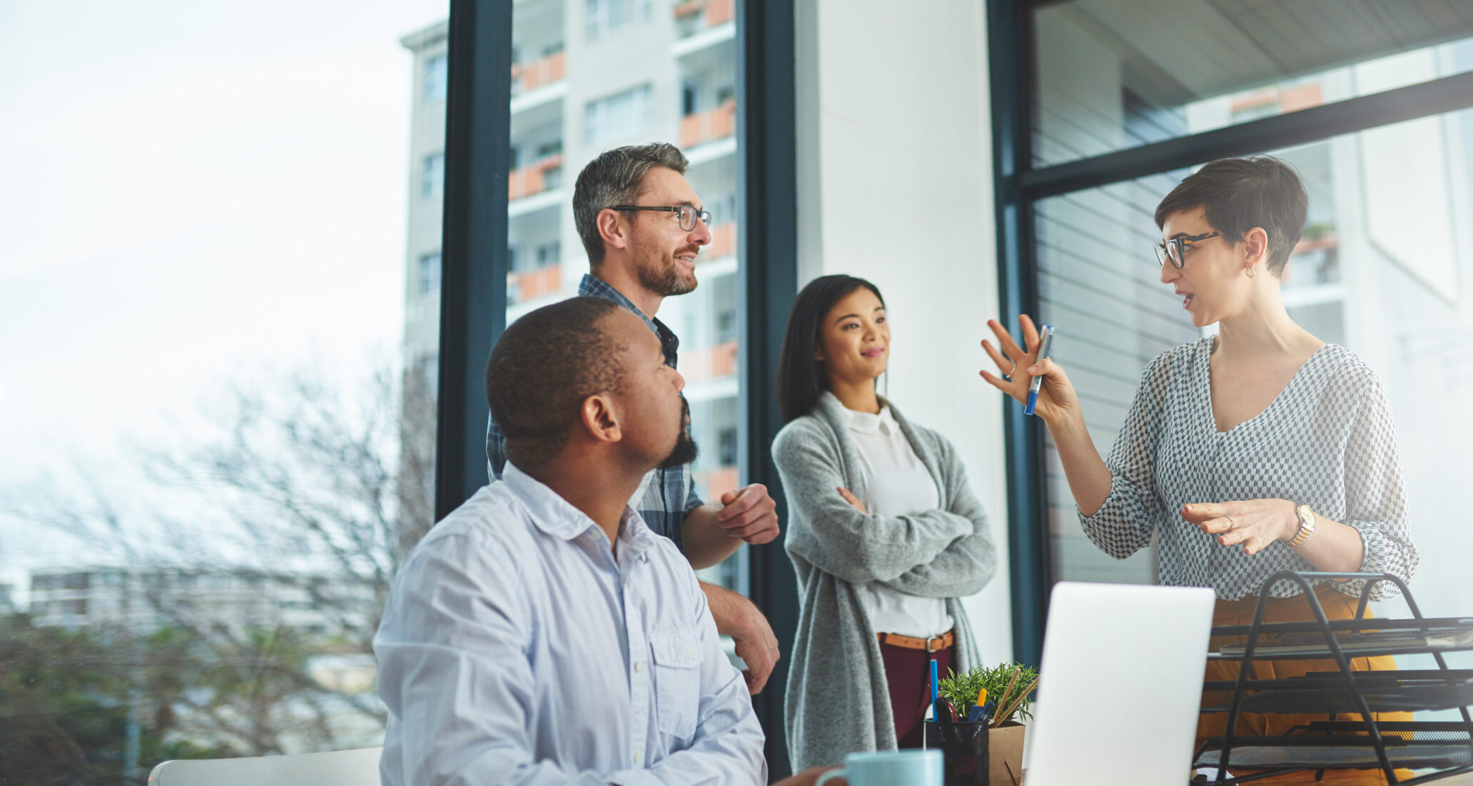Colleagues in discussion near window, one gesturing with pen, others listening attentively